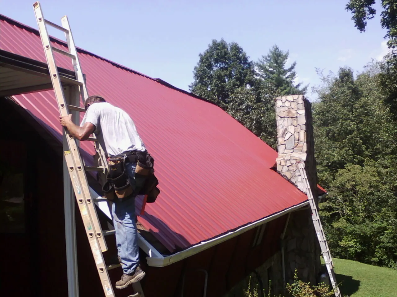 Roofer on a ladder beside a red metal roof and stone chimney — Soffit & Fascia Repair by Roofing Brothers in Painesville